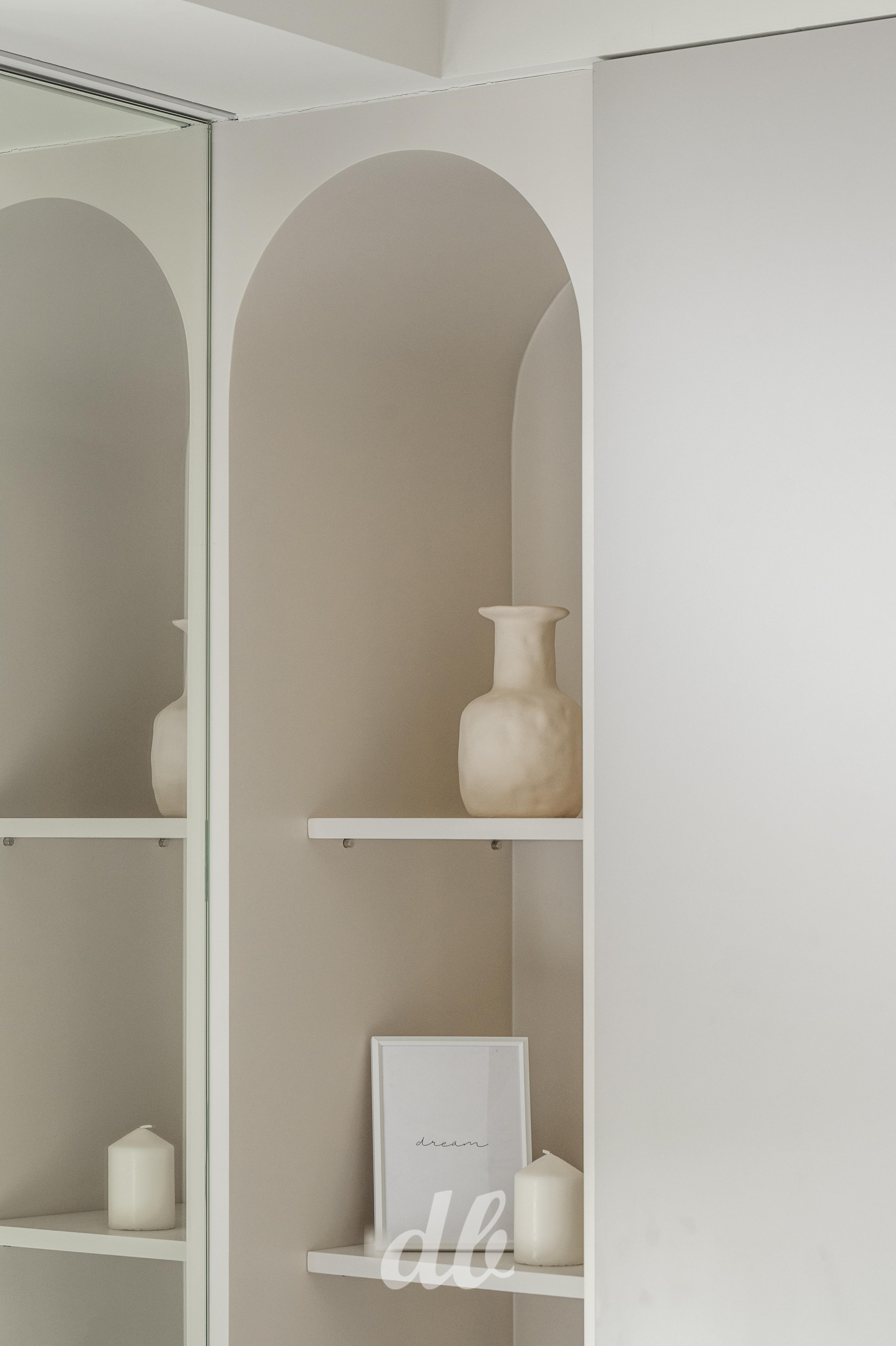 韓式奶油風壁龕細節：臥室牆面嵌入優雅的圓拱形壁龕設計，以簡約白色層板搭配米色系藝術陶瓶與蠟燭軟裝，透過圓潤流暢的曲線語彙取代銳利邊角，營造出極具層次感且純淨溫潤的居家視覺端景。