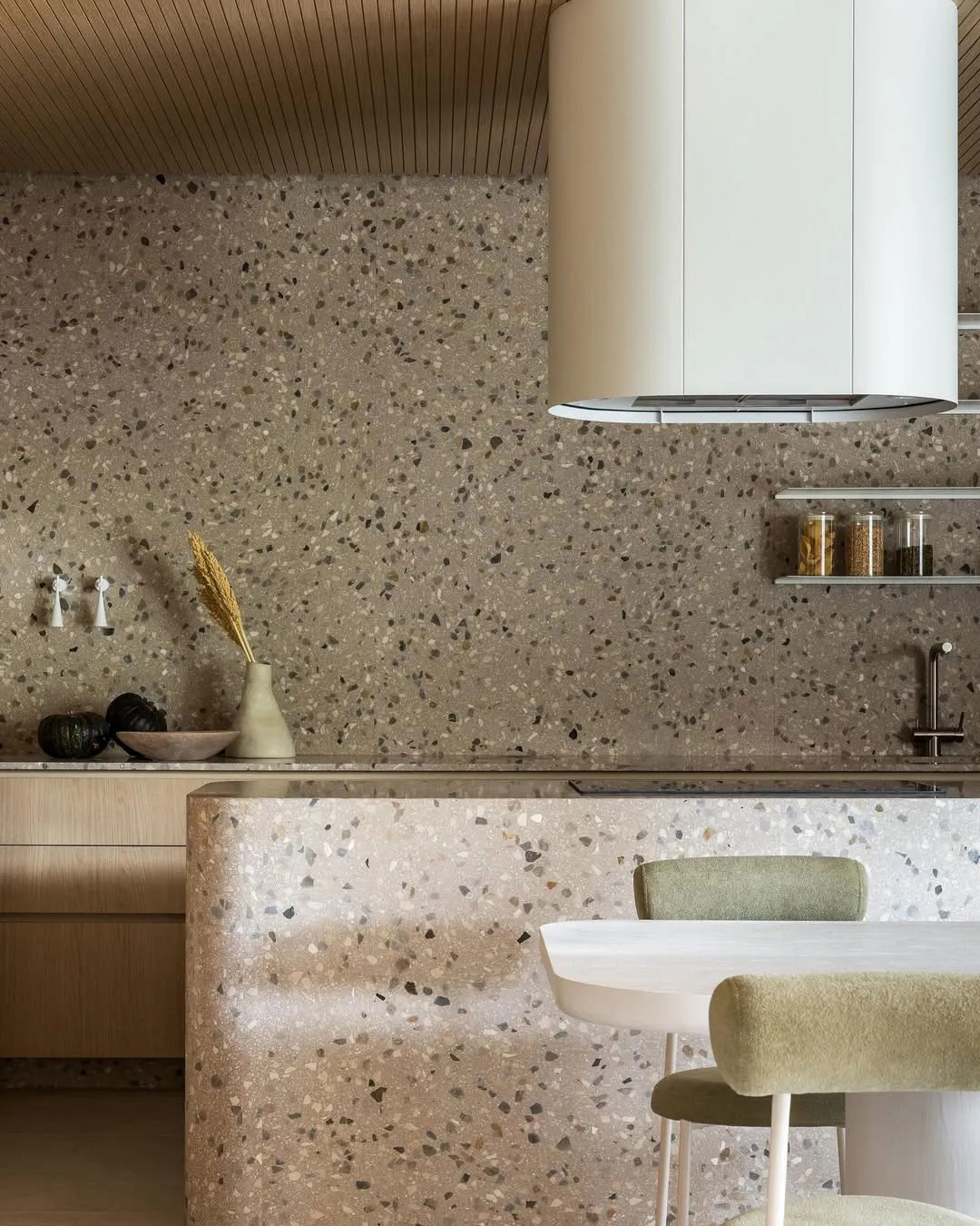 水磨石中島設計案例：圓弧導角的大面積暖灰色水磨石中島，搭配同材質通鋪牆面與白色極簡餐桌，在原木天花板的映襯下，展現異材質融合的自然美學與純粹質感。